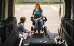 Eine Frau sitzt in einem elektrischen Rollstuhl und fährt eine Rampe in ein größeres Auto hoch. Eine weitere Frau ist neben der Rampe behilflich.