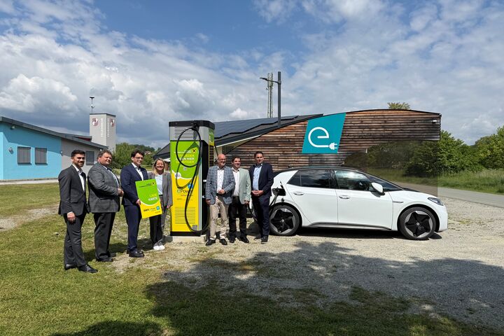 Eine Gruppe von Menschen steht im Freien um eine Ladestation. Daneben steht ein weißes Auto.