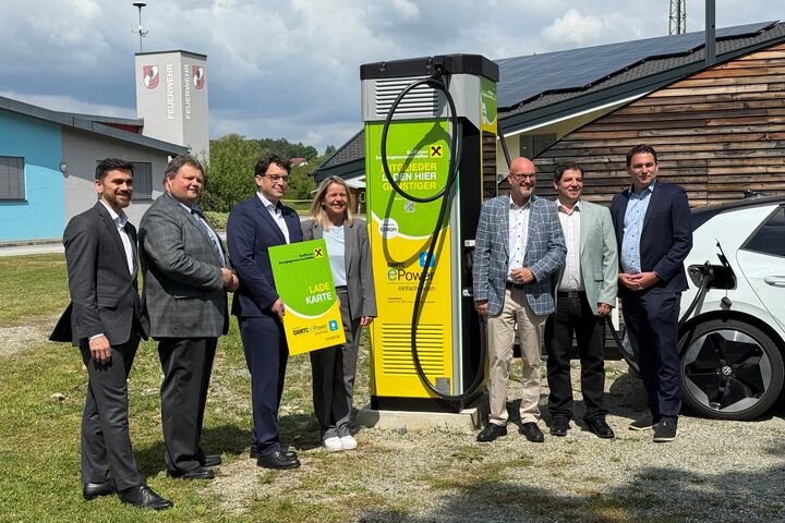 Eine Gruppe von Menschen steht im Freien um eine Ladestation daneben steht ein weißes Auto.