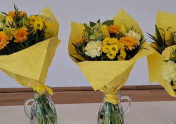 Das Foto zeigt fünf Blumensträuße mit gelbem Geschenkpapier umwickelt. Die Blumen selbst sind ebenfalls gelb und orange.