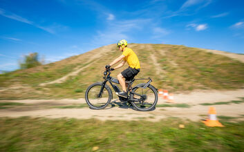 Der auto touring- Redakteur fährt mit einem E-Bike bei sommerlichem Wetter auf einem hügeligen Feldweg. An der Seite des Weges sind Pylonen aufgestellt.