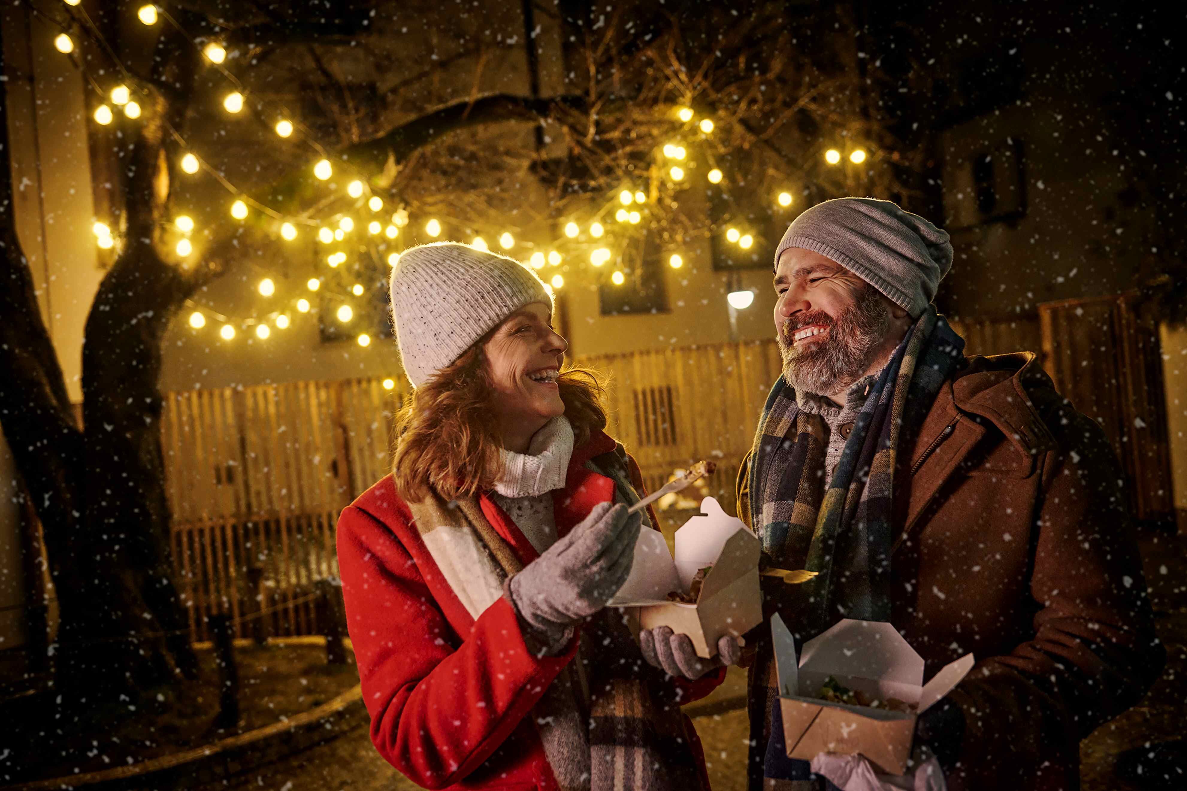 Ein lächelndes Paar isst bei winterlichem Wetter unter festlichen Lichterketten.
