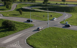 Eine Kreuzung mit Ampeln inmitten eines Verkehrsübungsplatzes. Neben den asphaltierten Straßen erstrecken sich grüne Wiesen. An der Kreuzung stehen vier Autos wartend vor der Ampel. Im Hintergrund ist eine Serpentinenstraße mit engen Kurven.