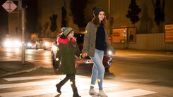 Ein Kind uns eine erwachsene Person überqueren in der Dunkelheit einen Zebrastreifen, im Hintergrund sieht man ein Auto.