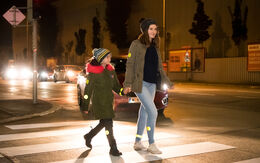 Ein Kind uns eine erwachsene Person überqueren in der Dunkelheit einen Zebrastreifen, im Hintergrund sieht man ein Auto.