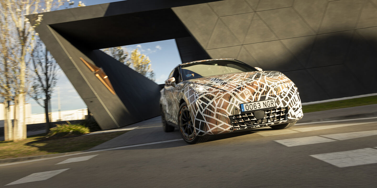 Ein mit Tarnfolie folierter Cupra Raval fährt bei einem futuristischen Gebäude vorbei.