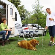 Auf einem Campingplatz sitzt eine Frau vor einem Wohnwagen, während ein Mann mit einem Hund auf der Wiese spielt.