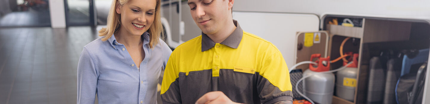 ÖAMTC Techniker erklärt einem weiblichen Mitglied etwas auf dem Tablet. Im Hintergrund sind die Gasanlage und Gasflaschen zu sehen