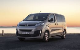 Grauer Citroen e-Jumpy, Ansicht seitlich von vorne, steht auf einer Straße vor einem Wasser. Im Hintergrund der Sonnenuntergang.