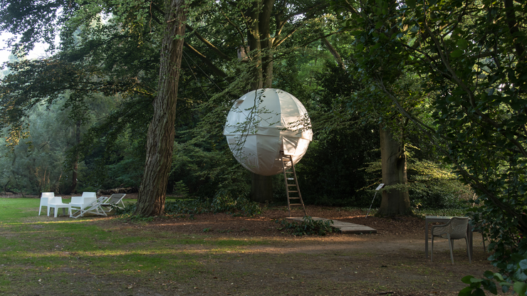 Eine runde, weiße Baumhauskugel hängt zwischen Bäumen im Wald, mit einer Leiter und Gartenstühlen in der Nähe.