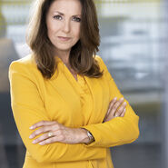 Porträtfoto einer Frau mit brünetten Haaren und verschränkten Armen, die in einem gelben Blazer vor einer Glaswand steht