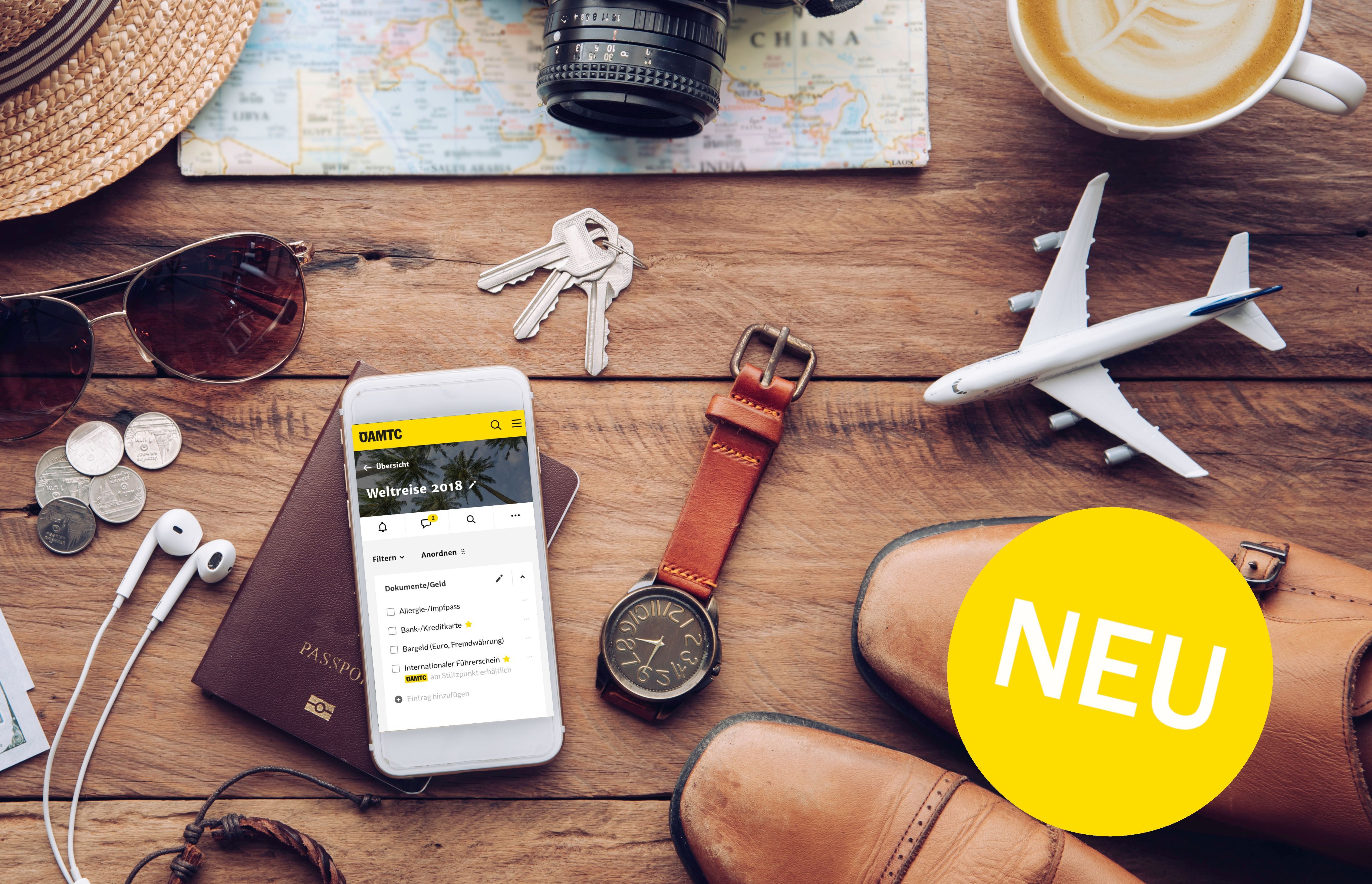 Reiseutensilien auf einem Tisch: Smartphone mit ÖAMTC-App, Uhr, Schlüssel, Sonnenbrille, Kamera, Modellflugzeug und Tasse Kaffee. Ein gelber Kreis mit "NEU" ist sichtbar.