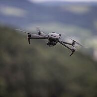 Drohne_ÖAMTC_1 Auf dem Bild sieht man eine graue Drohne mit vier Rotoren im Flug, im Hintergrund verschwommene Landschaft.