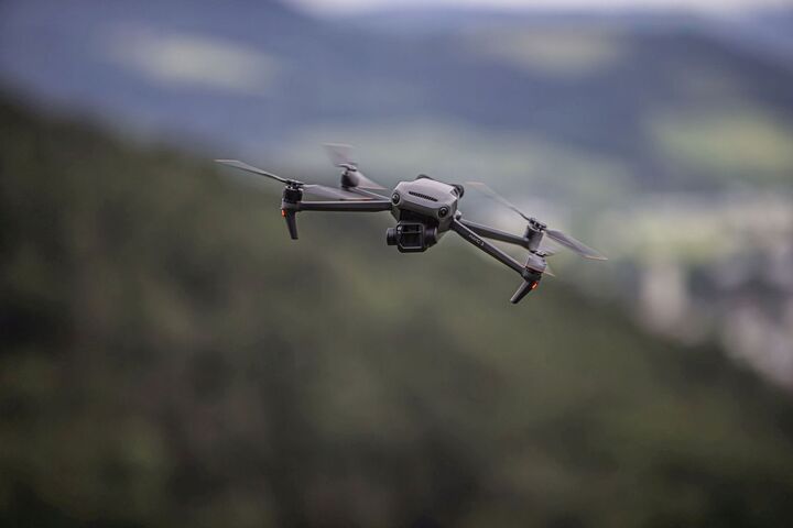 Auf dem Bild sieht man eine graue Drohne mit vier Rotoren im Flug, im Hintergrund verschwommene Landschaft.