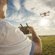 Auf dem Bild sieht man einen Teil des Oberkörpers und Kopfes eines Mannes. Er trägt ein weißes Hemd und hält eine Fernsteuerung in den Händen. Am Himmel sieht man eine Drohen, die von diesem Mann gesteuert wird.