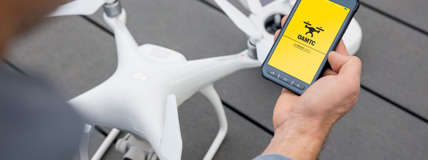 Eine weiße Drohne steht auf einem grauen Boden. im Vordergrund ist eine Person sichtbar, diese hält in der rechten Hand ein Smartphone, am Display ist die ÖAMTC Drohnen App geöffnet.