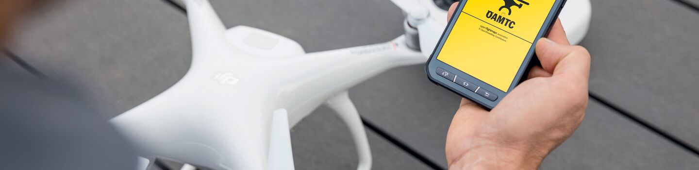 Eine weiße Drohne steht auf einem grauen Boden. im Vordergrund ist eine Person sichtbar, diese hält in der rechten Hand ein Smartphone, am Display ist die ÖAMTC Drohnen App geöffnet.