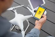 Eine weiße Drohne steht auf einem grauen Boden. im Vordergrund ist eine Person sichtbar, diese hält in der rechten Hand ein Smartphone, am Display ist die ÖAMTC Drohnen App geöffnet.