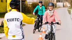 Zwei ältere Personen üben mit dem E-Bike Kurvenfahren. Im Vordergrund gibt die ÖAMTC-Instruktorin Anweisungen.