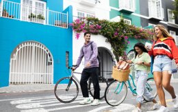 3 Personen gehen auf einer Straße und lächeln. Zwei schieben ein Fahrrad. Im Hintergrund sind blau-türkise Häuser.