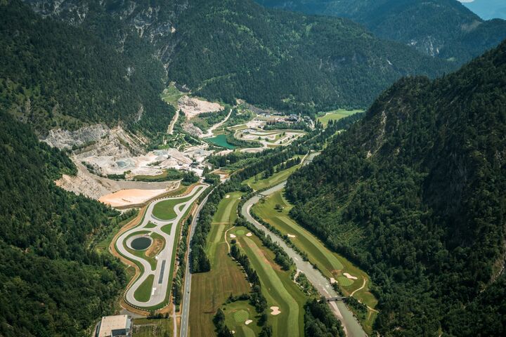 Luftaufnahme der Experience World Saalfelden - man sieht eine asphaltierte Rundstrecke, einen Offroad-Bereich, einen See und Teile eines Golfplatzes, mitten im Tal eines malerischen Alpenpanoramas