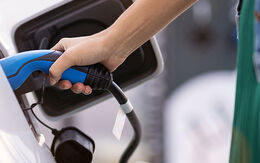 Eine Frau lädt ein Elektroauto. Der blaue Ladestecke wird mit der rechten Hand gehalten.