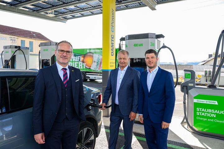 Auf dem Bild sind drei Männer in Anzügen zu sehen, die neben der neuen Ladestation stehen. Links im Bild ist Harald Großauer (Landesdirektor ÖAMTC Oberösterreich), er trägt einen dunklen Anzug mit Rot-schwarz-gestreifter Krawatte und eine Brille. Etwas weiter rechts im Bild stehen Franz Ortner und Michael Ortner (Eigentümer und Geschäftsführer Autohaus Ortner), beide tragen dunkelblaue Anzüge und hellblaue Hemden. Franz Ortner ist gerade dabei das Elektorauto aufzuladen.