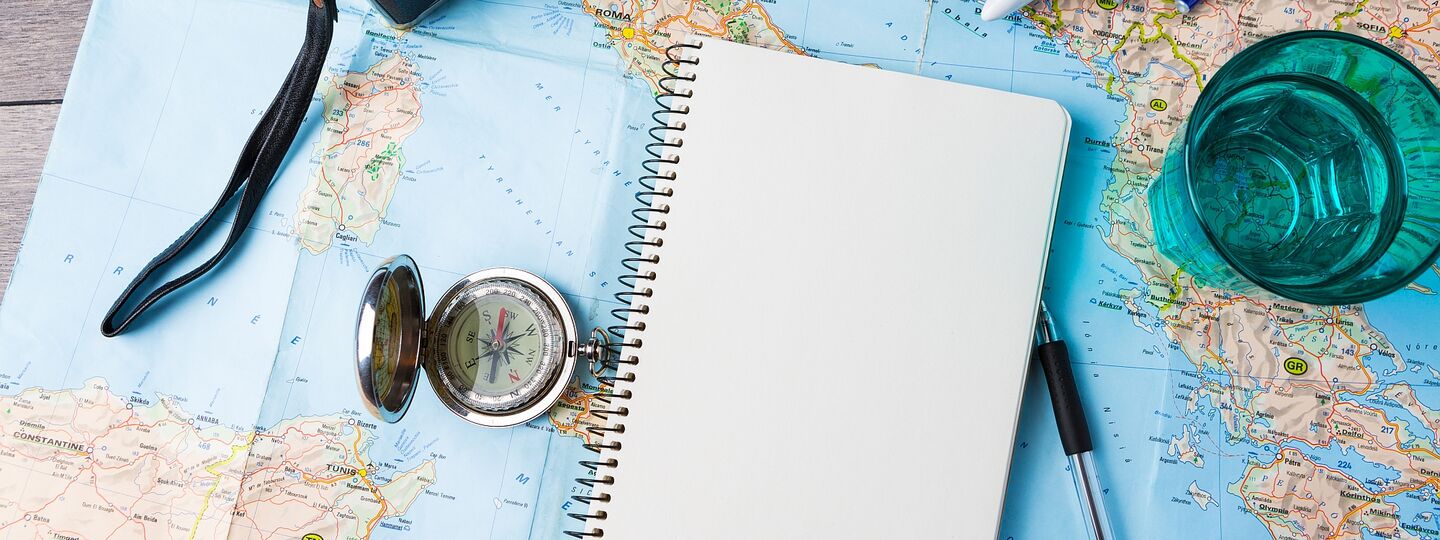 Eine Europakarte liegt auf einem Holztisch. Auf der Karte liegt ein Kompass, eine Kamera, ein Notizbuch mit einem Kugelschreiber, ein blaues Wasserglas und ein Modelflugzeug.