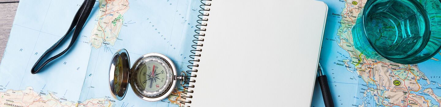 Eine Europakarte liegt auf einem Holztisch. Auf der Karte liegt ein Kompass, eine Kamera, ein Notizbuch mit einem Kugelschreiber, ein blaues Wasserglas und ein Modelflugzeug.