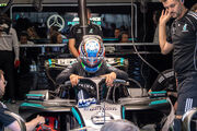Ein Formel-1-Fahrer in voller Montur mit Rennanzug und -helm steht neben seinem Formel 1 Auto, um ihn herum arbeiten viele Teammitglieder.