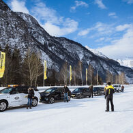 Im Bild sind fünf Autos zu sehen, die am Rand einer Schneefahrbahn parken. Die Lenkerinnen und Lenker steigen aus und schauen zu einem Fahrtechnikinstruktor. Im Hintergrund sind Berge und Wälder zu sehen.