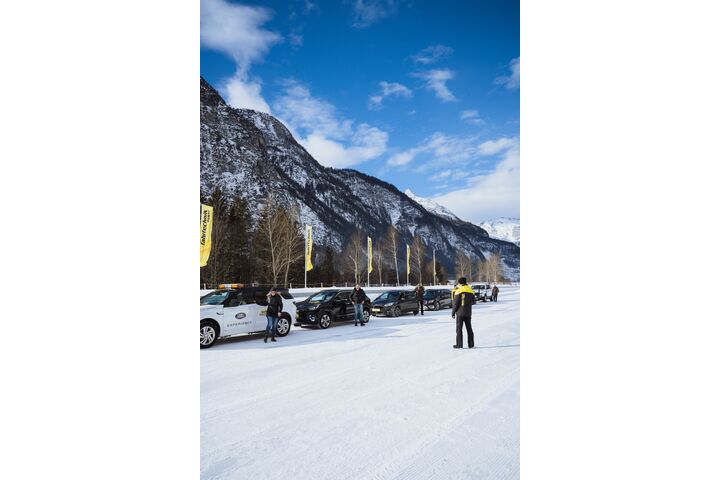 Im Bild sind fünf Autos zu sehen, die am Rand einer Schneefahrbahn parken. Die Lenkerinnen und Lenker steigen aus und schauen zu einem Fahrtechnikinstruktor. Im Hintergrund sind Berge und Wälder zu sehen.