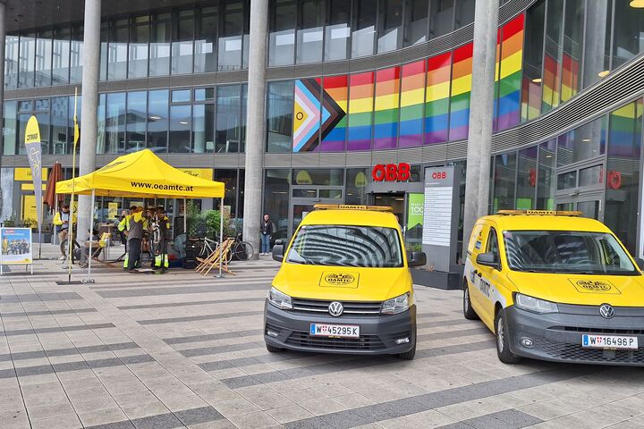Fahrradfahrer:innen lassen sich ihre Fahrränder am ÖAMTC PopUp Fahrrad-Stützpunkt vor dem ÖBB Hauptbahnhof Wien reparieren, vor dem Gebäude sind zwei gelbe Pannenautos und der PopUp Stand zu sehen