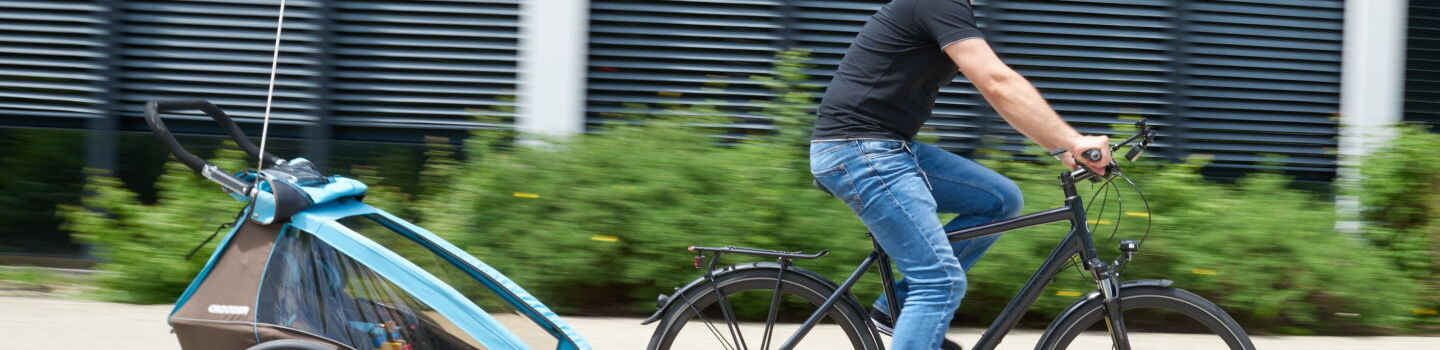 Ein Mann fährt mit einem Fahrrad auf einem gepflasterten Weg. Er trägt einen türkisen Helm, ein schwarzes T-Shirt und blaue Jeans. An seinem Fahrrad ist ein Kinderfahrradanhänger befestigt, in dem sich ein Kind befindet.