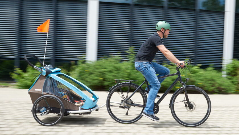 Ein Mann fährt mit einem Fahrrad auf einem gepflasterten Weg. Er trägt einen türkisen Helm, ein schwarzes T-Shirt und blaue Jeans. An seinem Fahrrad ist ein Kinderfahrradanhänger befestigt, in dem sich ein Kind befindet.