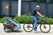 Ein Mann fährt mit einem Fahrrad auf einem gepflasterten Weg. Er trägt einen türkisen Helm, ein schwarzes T-Shirt und blaue Jeans. An seinem Fahrrad ist ein Kinderfahrradanhänger befestigt, in dem sich ein Kind befindet.