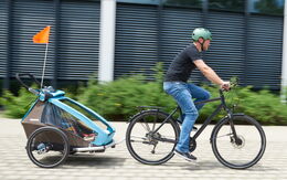 Ein Mann fährt mit einem Fahrrad auf einem gepflasterten Weg. Er trägt einen türkisen Helm, ein schwarzes T-Shirt und blaue Jeans. An seinem Fahrrad ist ein Kinderfahrradanhänger befestigt, in dem sich ein Kind befindet.