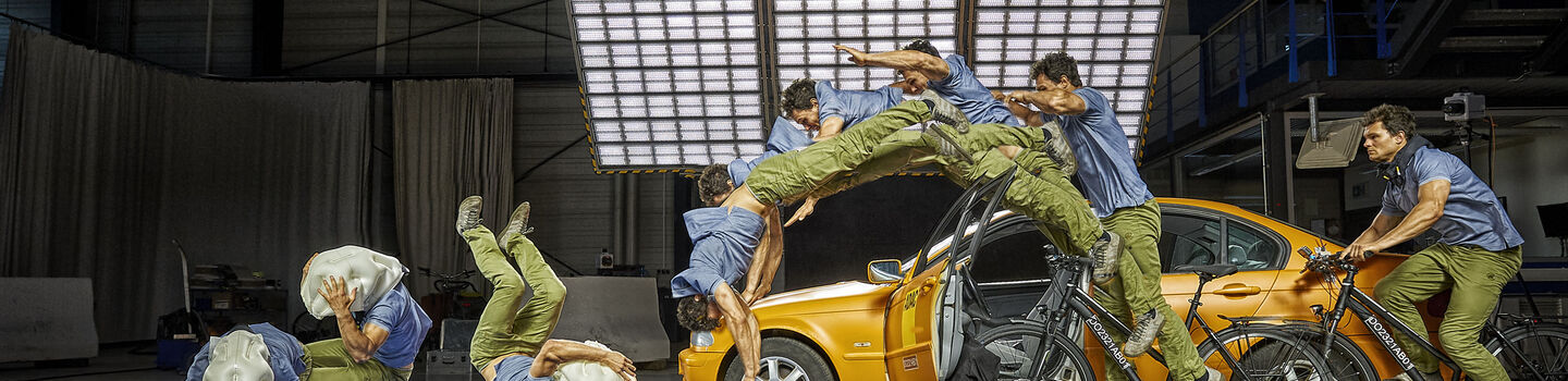 Mehrere Bilder eines Mannes sind in einer Bildsequenz zusammengefügt, die einen Stunt zeigt. Der Mann trägt ein blaues Hemd und grüne Hosen und stürzt von einem Fahrrad, das mit einem orangefarbenen Auto kollidiert ist. Die Sequenz zeigt ihn in verschiedenen Stadien des Sturzes, beginnend vom Moment des Aufpralls bis zum Aufprall auf eine blaue Matte auf dem Boden. Die Szene findet in einer großen Halle mit professioneller Beleuchtung statt.