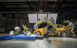 Mehrere Bilder eines Mannes sind in einer Bildsequenz zusammengefügt, die einen Stunt zeigt. Der Mann trägt ein blaues Hemd und grüne Hosen und stürzt von einem Fahrrad, das mit einem orangefarbenen Auto kollidiert ist. Die Sequenz zeigt ihn in verschiedenen Stadien des Sturzes, beginnend vom Moment des Aufpralls bis zum Aufprall auf eine blaue Matte auf dem Boden. Die Szene findet in einer großen Halle mit professioneller Beleuchtung statt.
