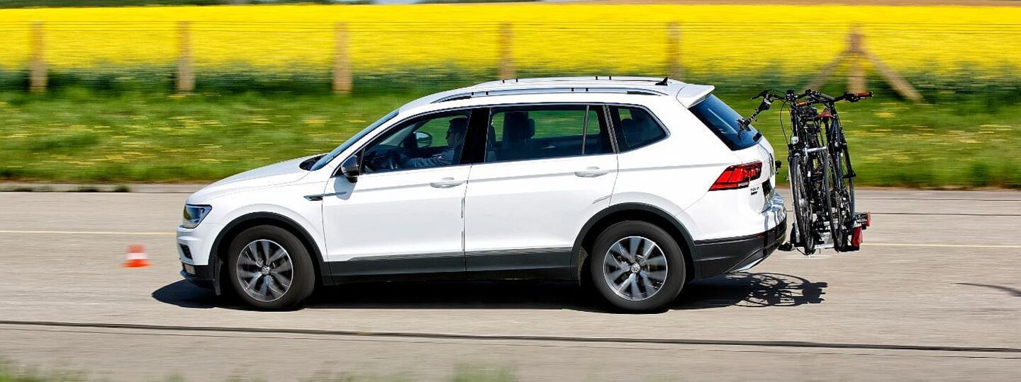 Weißer SUV fährt auf einer Straße fährt. Am Heck des Fahrzeugs ist ein Fahrradträger mit zwei montierten Fahrrädern befestigt. Im Hintergrund erstreckt sich ein Rapsfeld, der Himmel ist blau.