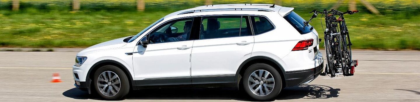 Weißer SUV fährt auf einer Straße fährt. Am Heck des Fahrzeugs ist ein Fahrradträger mit zwei montierten Fahrrädern befestigt. Im Hintergrund erstreckt sich ein Rapsfeld, der Himmel ist blau.