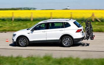 Weißer SUV fährt auf einer Straße fährt. Am Heck des Fahrzeugs ist ein Fahrradträger mit zwei montierten Fahrrädern befestigt. Im Hintergrund erstreckt sich ein Rapsfeld, der Himmel ist blau.