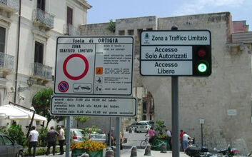 Zwei Schilder in einer italienischen Stadt. Das linke Schild weist auf eine Zone mit beschränktem Verkehr (Zona Traffico Limitato) in Ortigia hin, mit einem Durchfahrtsverbotsschild. Das rechte Schild hat eine Ampel und die Aufschriften "Accesso Solo Autorizzati" und "Accesso Libero". Im Hintergrund sind Gebäude, Autos und Menschen zu sehen.