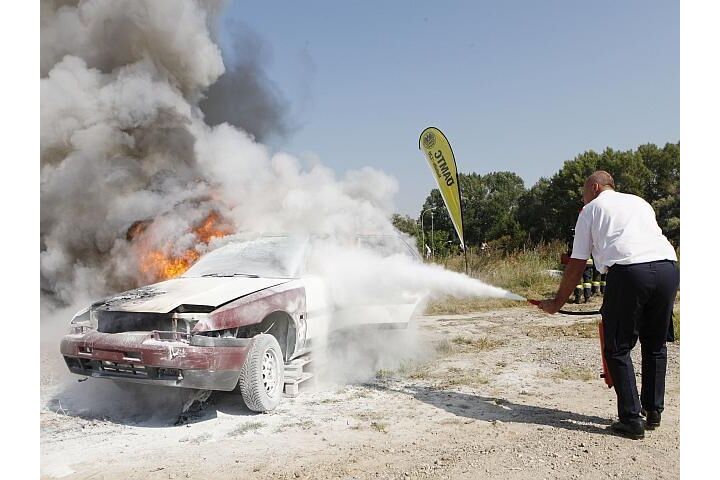 Auf einer Schotterfläche steht ein verbranntes Fahrzeug, ein Mann löscht dieses mit einem Handfeuerlöscher. Im Hintergrund sind eine ÖAMTC Fahrtechnik Fahne und Bäume.