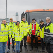 Gruppenfoto mit zwei Instruktoren der Fahrtechnik gemeinsam mit 5 Busfahrer:innen des Flughafen Wiens vor Flughafen-Kulisse