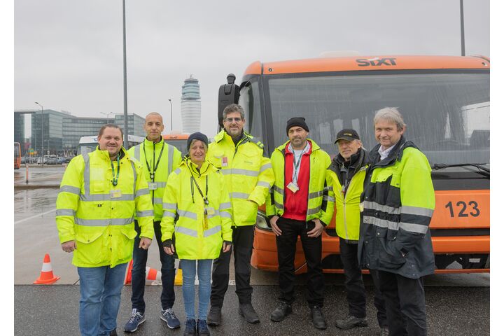 Gruppenfoto mit zwei Instruktoren der Fahrtechnik gemeinsam mit 5 Busfahrer:innen des Flughafen Wiens vor Flughafen-Kulisse