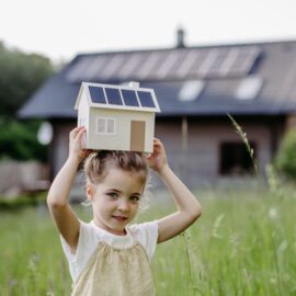 Ein Kleinkind in beiger Sommerkleidung steht auf einer grünen Blumenwiese und hält ein Miniaturmodell von einem Haus am Kopf. Am Dach ist eine Photovoltaikanlage montiert.