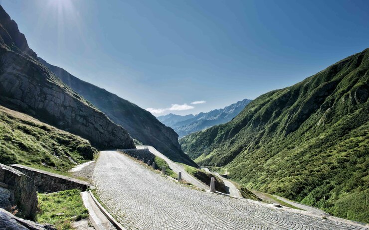 Das Foto zeigt die geplasterte Passtraße über den Gotthard in der Schweiz bei strahlendem blauen Himmel und mit Bergen zu beiden Seiten.