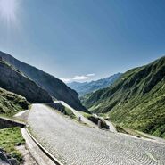 Das Foto zeigt die geplasterte Passtraße über den Gotthard in der Schweiz bei strahlendem blauen Himmel und mit Bergen zu beiden Seiten.