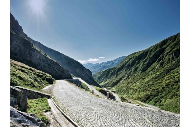 Das Foto zeigt die geplasterte Passtraße über den Gotthard in der Schweiz bei strahlendem blauen Himmel und mit Bergen zu beiden Seiten.